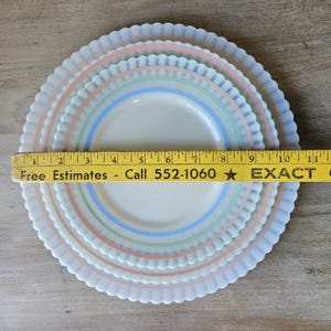 May include: Set of three white plates with pastel pink, green, and blue stripes. The plates have a scalloped edge and are arranged in a nested configuration. A ruler is placed across the plates. The text on the ruler says "Free Estimates - Call 552-1060".