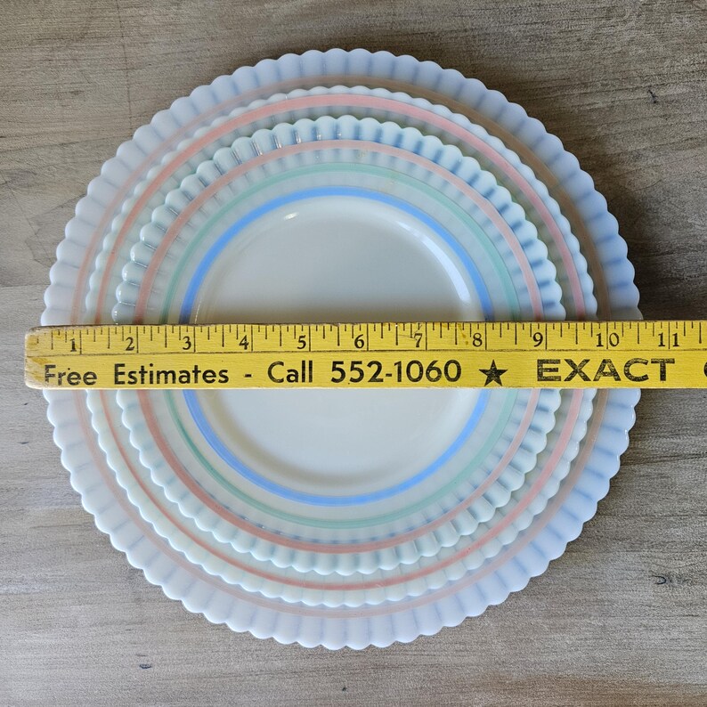 May include: Set of three white plates with pastel pink, green, and blue stripes. The plates have a scalloped edge and are arranged in a nested configuration. A ruler is placed across the plates. The text on the ruler says "Free Estimates - Call 552-1060".