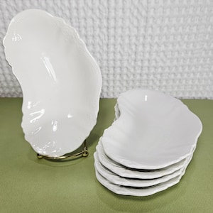 May include: A set of white, oyster-shaped serving dishes. One dish is displayed on a gold-colored stand, while a stack of similar dishes rests nearby. The dishes have a textured surface and a scalloped edge, suggesting a coastal or nautical theme.