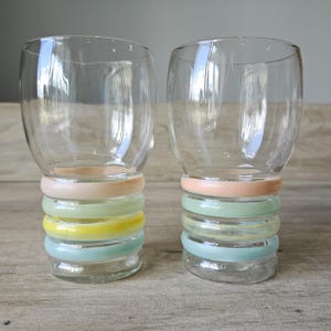 Pode incluir: Dois copos de vidro transparente com um design de an&eacute;is coloridos empilhados na base. Os an&eacute;is s&atilde;o tons pastel de rosa, amarelo, verde e azul. Os copos t&ecirc;m um formato arredondado.