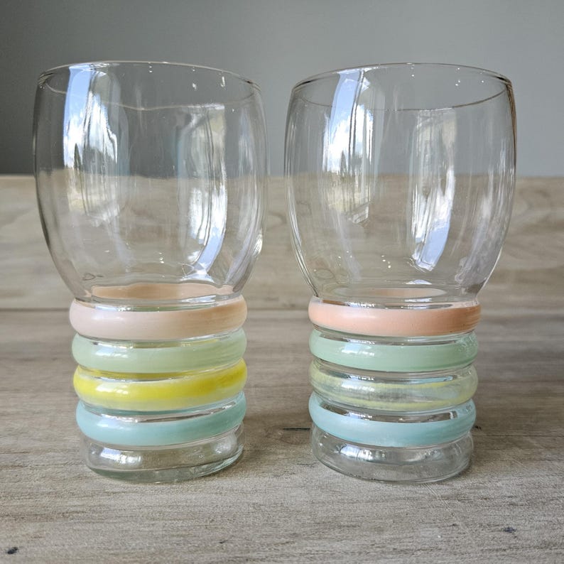 Pode incluir: Dois copos de vidro transparente com um design de an&eacute;is coloridos empilhados na base. Os an&eacute;is s&atilde;o tons pastel de rosa, amarelo, verde e azul. Os copos t&ecirc;m um formato arredondado.