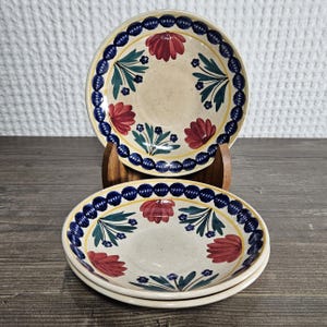May include: A set of three ceramic plates with a cream-colored base. Each plate features a blue beaded border, a yellow inner border, and red floral designs with green leaves. The plates are stacked on a wooden surface.
