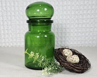 May include: A cylindrical green glass apothecary jar with a matching rounded lid. Beside the jar sits a small, dark brown woven twig nest containing two speckled, off-white eggs, accompanied by a sprig of small white flowers.