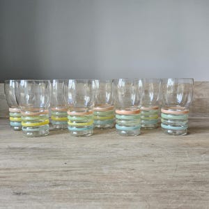 Pode incluir: Um conjunto de copos transparentes com um design de faixas em cores pastel. Cada copo apresenta uma pilha de faixas horizontais em tons de rosa, amarelo, verde e azul. Os copos est&atilde;o dispostos sobre uma superf&iacute;cie de madeira clara.