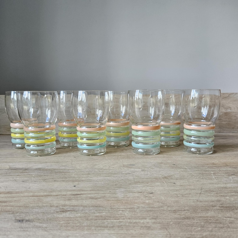 Pode incluir: Um conjunto de copos transparentes com um design de faixas em cores pastel. Cada copo apresenta uma pilha de faixas horizontais em tons de rosa, amarelo, verde e azul. Os copos est&atilde;o dispostos sobre uma superf&iacute;cie de madeira clara.