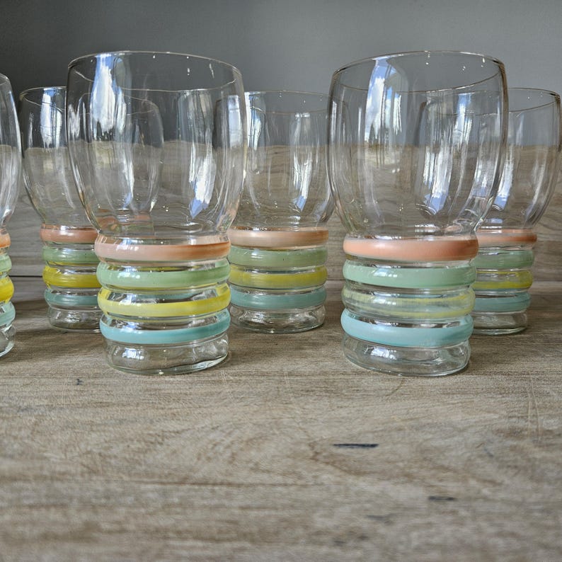 Pode incluir: Um conjunto de copos de vidro transparente com um design de an&eacute;is coloridos empilhados na base. Os an&eacute;is s&atilde;o em tons pastel de rosa, verde, amarelo e azul. Os copos est&atilde;o sobre uma superf&iacute;cie de madeira.