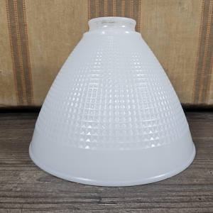 May include: A white glass lampshade with a textured, geometric pattern. The shade has a conical shape with a wide base and a narrower top. The surface is covered in a repeating pattern of squares and triangles.