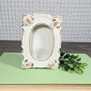 Pode incluir: Moldura de cerâmica branca com abertura oval, adornada com detalhes de rosas cor-de-rosa e acabamentos dourados. A moldura tem um design decorativo e ornamentado. Um ramo de folhas verdes repousa ao lado da moldura.