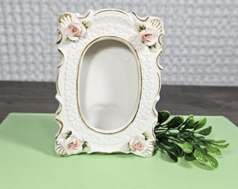 Puede incluir: Marco de fotos de cerámica blanca con una abertura ovalada, adornado con acentos de rosas rosas y ribetes dorados. El marco tiene un diseño decorativo y ornamentado. Una ramita de hojas verdes descansa junto al marco.