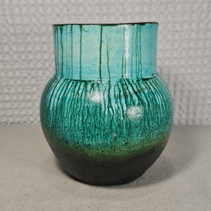 May include: A handcrafted ceramic vase with a rounded body and a wide opening. The vase features a turquoise top with vertical green streaks that fade into a darker green and black base. The vase is a decorative home accent.
