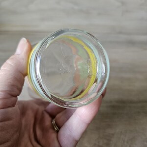 Pode incluir: Copo de vidro transparente com um design colorido. O copo tem uma faixa amarela, verde e p&ecirc;ssego ao redor da parte superior. O copo &eacute; segurado por uma m&atilde;o.