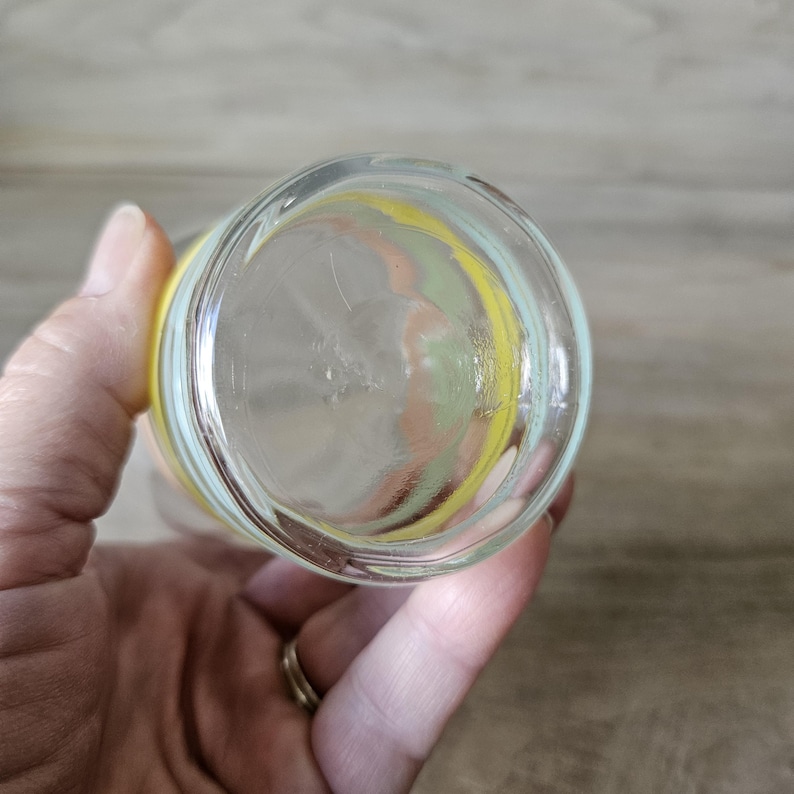 Pode incluir: Copo de vidro transparente com um design colorido. O copo tem uma faixa amarela, verde e p&ecirc;ssego ao redor da parte superior. O copo &eacute; segurado por uma m&atilde;o.