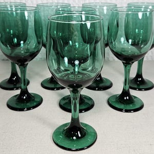 May include: A set of seven matching emerald green glass wine goblets. Each glass features a smooth, rounded bowl, a slender stem, and a circular base. The glassware is uniform in color and design, showcasing a deep, translucent green hue throughout.