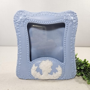 May include: A light blue, ornate picture frame with a rectangular opening. The frame features a raised floral and beaded design. A white cameo with a woman's profile is centered at the bottom. The frame is made of a solid material.