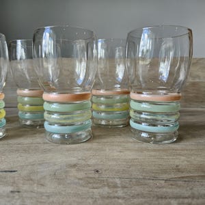Pode incluir: Um conjunto de seis copos de vidro transparente com um design colorido de an&eacute;is empilhados na base. Os an&eacute;is s&atilde;o tons pastel de rosa, verde, amarelo e azul. Os copos est&atilde;o dispostos sobre uma superf&iacute;cie de madeira.