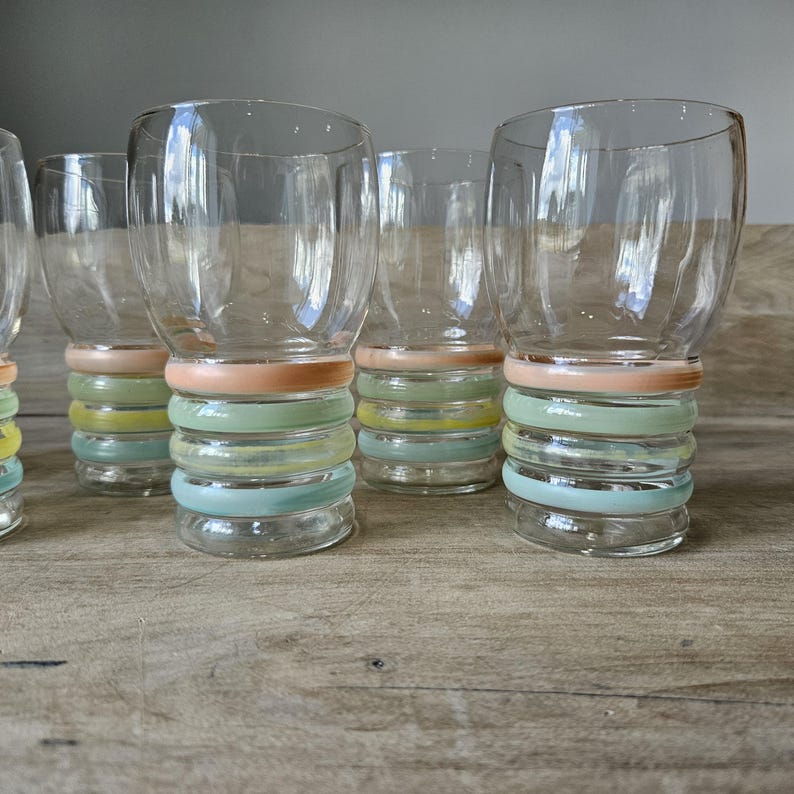 Pode incluir: Um conjunto de seis copos de vidro transparente com um design colorido de an&eacute;is empilhados na base. Os an&eacute;is s&atilde;o tons pastel de rosa, verde, amarelo e azul. Os copos est&atilde;o dispostos sobre uma superf&iacute;cie de madeira.