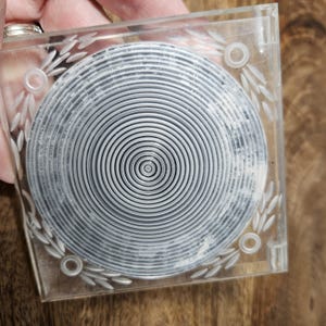 May include: A clear plastic coaster with a circular design of concentric circles in the center. The coaster has a decorative border of leaves around the edge.