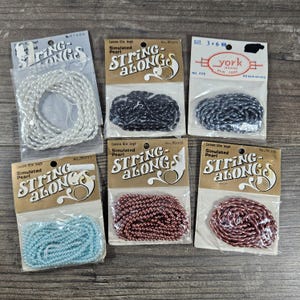 May include: A collection of vintage simulated pearl string beads in various colors, including white, black, blue, and shades of red. The beads are packaged in clear plastic with retro-style cardboard labels. The labels read "String Along".