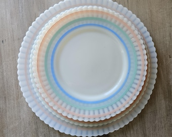 May include: A stack of white, scalloped-edge plates with pastel-colored rings. The plates are arranged in a nested fashion, with the largest plate at the bottom and the smallest at the top. The rings are in shades of peach, green, and blue.