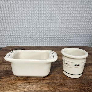 May include: Two off-white ceramic containers with a festive design. One is a rectangular dish with small handles, and the other is a small cylindrical pot. Both have a green and red holly leaf pattern around the edges. They sit on a wooden surface.