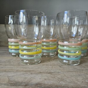 Pode incluir: Conjunto de copos transparentes com um design de faixas em cores pastel. Cada copo apresenta uma s&eacute;rie de faixas horizontais em tons de rosa, amarelo, verde e azul. Os copos est&atilde;o dispostos sobre uma superf&iacute;cie de madeira.