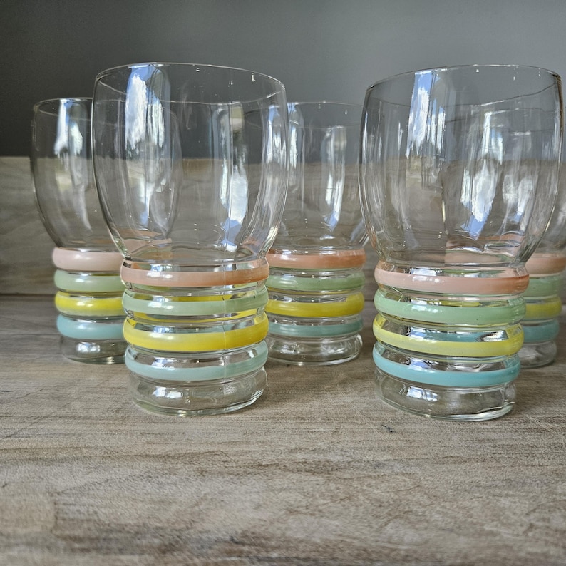 Pode incluir: Conjunto de copos transparentes com um design de faixas em cores pastel. Cada copo apresenta uma s&eacute;rie de faixas horizontais em tons de rosa, amarelo, verde e azul. Os copos est&atilde;o dispostos sobre uma superf&iacute;cie de madeira.