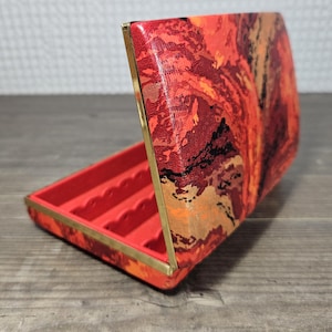 May include: Open red jewelry box with a vibrant, abstract pattern in shades of red, orange, and gold. The box has a red interior with several compartments and gold-colored metal trim. The box is open, revealing the interior.