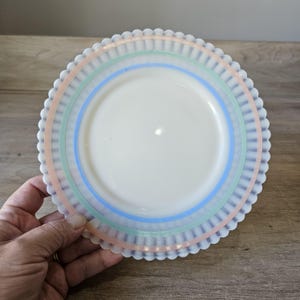 May include: A white plate with a scalloped edge and pastel stripes in pink, green, and blue. The plate has a smooth, glossy surface and a decorative design. This vintage plate is perfect for serving food.