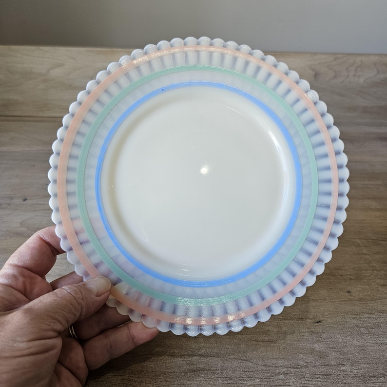 May include: A white plate with a scalloped edge and pastel stripes in pink, green, and blue. The plate has a smooth, glossy surface and a decorative design. This vintage plate is perfect for serving food.