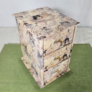 May include: A small wooden box with three drawers, decorated with a floral pattern featuring angels and roses. The drawers have gold-colored handles.