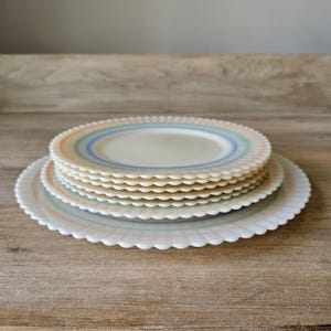 May include: A stack of white plates with scalloped edges and pastel-colored stripes. The plates are arranged on a wooden surface, showcasing a vintage aesthetic. The plates have a retro design with a light blue, green, and orange color scheme.
