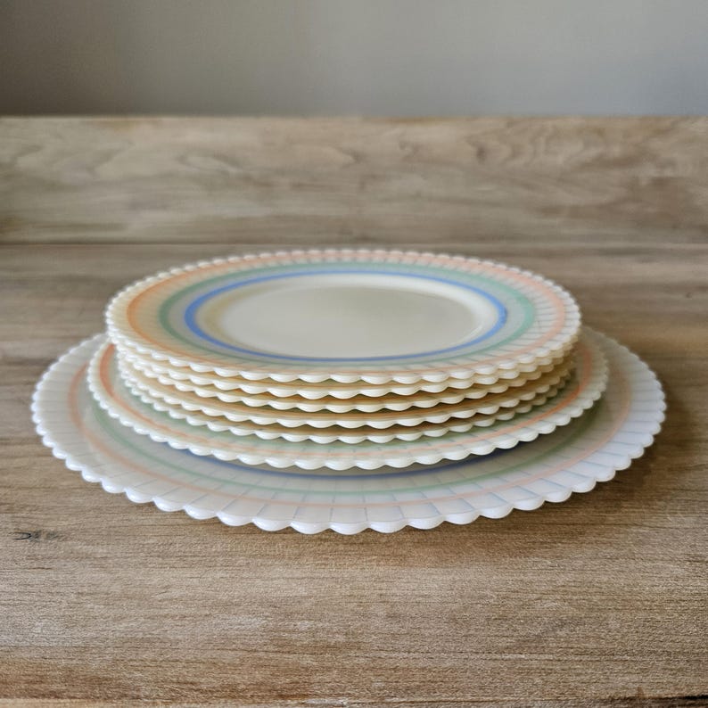 May include: A stack of white plates with scalloped edges and pastel-colored stripes. The plates are arranged on a wooden surface, showcasing a vintage aesthetic. The plates have a retro design with a light blue, green, and orange color scheme.
