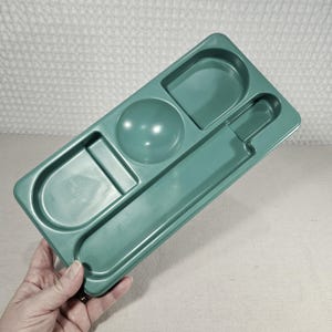 Pode incluir: Uma bandeja de plástico verde-azulado com vários compartimentos. A bandeja tem uma seção arredondada, um semicírculo e uma seção retangular longa. A bandeja é segurada por uma mão, e o fundo é uma superfície texturizada esbranquiçada.