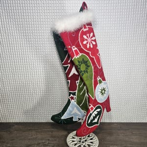 May include: Two Christmas stockings, one red and one green, with white faux fur trim. The red stocking features white snowflake and ornament designs. The green stocking has a Christmas tree design. Both stockings are displayed on a white snowflake-shaped stand.