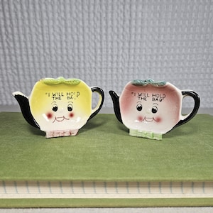 May include: Two ceramic teapot-shaped trinket holders. One is yellow with a black handle and spout, the other is pink with a black handle and spout. Both have a smiling face and the text "I WILL HOLD THE BAG".