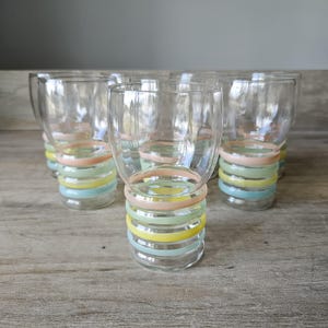 Pode incluir: Um conjunto de seis copos transparentes com um design de an&eacute;is pastel na base. Os an&eacute;is s&atilde;o em tons de rosa, amarelo, verde e azul. Os copos est&atilde;o dispostos sobre uma superf&iacute;cie de madeira. Cada copo tem cerca de 10 cm de altura.