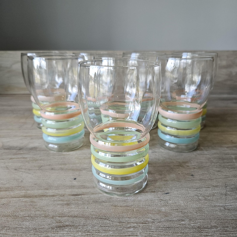 Pode incluir: Um conjunto de seis copos transparentes com um design de an&eacute;is pastel na base. Os an&eacute;is s&atilde;o em tons de rosa, amarelo, verde e azul. Os copos est&atilde;o dispostos sobre uma superf&iacute;cie de madeira. Cada copo tem cerca de 10 cm de altura.