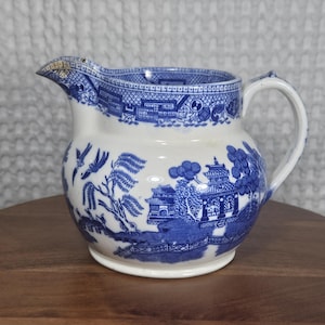 May include: A white ceramic creamer pitcher featuring a blue transferware pattern depicting a traditional willow landscape with pagodas, trees, and birds. The pitcher has a curved handle, a wide spout, and a decorative geometric border along the rim.