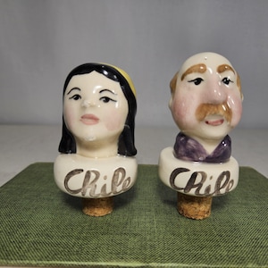 May include: A pair of ceramic bottle stoppers with painted faces. The stoppers are shaped like a woman and a man. Both stoppers have the word "Chile" written on the base.