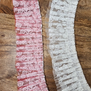 May include: Two pieces of lace trim. One is white, the other is pink with a red backing. Both trims have a floral pattern and are ruffled. The lace is on a wooden surface.