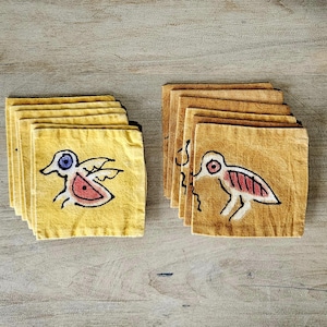 May include: Two stacks of square, yellow fabric coasters with hand-drawn bird designs. One stack features a bird with a red body, blue eye, and wings. The other stack has a bird with a red striped body and a red eye.