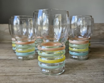 May include: Set of clear glass tumblers with colorful bands of pastel colors around the base. The bands include peach, green, yellow, and blue. The glasses are arranged on a wooden surface.
