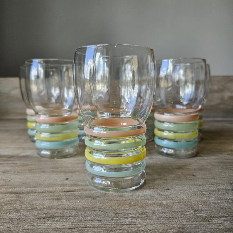 Pode incluir: Conjunto de copos de vidro transparente com faixas coloridas em tons pastel ao redor da base. As faixas incluem p&ecirc;ssego, verde, amarelo e azul. Os copos est&atilde;o dispostos sobre uma superf&iacute;cie de madeira.