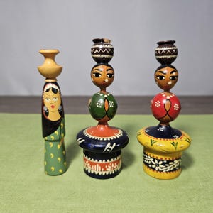 May include: Three handcrafted wooden figurines with painted details. The figurines feature stacked elements, including faces, spheres, and pot-like shapes, in colors like yellow, green, red, and blue. The central figurine has a face with large eyes.