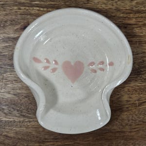 May include: Off-white ceramic spoon rest with a pink heart and leaf design. The spoon rest has a curved shape with a raised edge and a small indentation for the spoon handle. The speckled ceramic adds a rustic touch.