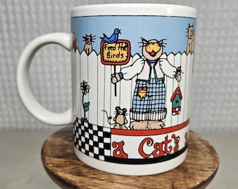 May include: White ceramic mug with a whimsical cat design. The mug features a cartoon cat holding a sign that reads "Feed the Birds." Other illustrations include a mouse, birdhouse, and a fence. The bottom of the mug reads "A Cat's".