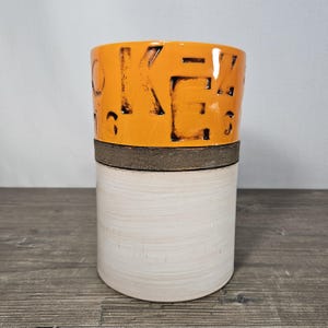 May include: A cylindrical ceramic vase with a two-tone design. The top section is bright orange with the word "OKEY" carved into it. The bottom section is a light beige color. A dark brown band separates the two sections. The vase is on a wooden surface.
