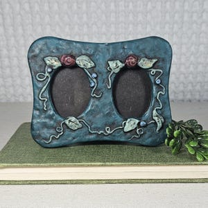 Pode incluir: Uma moldura para fotos dupla azul-petróleo com aberturas ovais. A moldura é decorada com rosas esculpidas, folhas e vinhas. A moldura está em um livro verde.