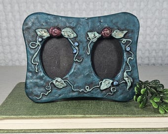 Pode incluir: Uma moldura para fotos dupla azul-petróleo com aberturas ovais. A moldura é decorada com rosas esculpidas, folhas e vinhas. A moldura está em um livro verde.