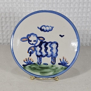 May include: A round ceramic plate featuring a hand-painted blue sheep standing on a green patch of grass. The design includes a blue cloud and a small bird above the sheep, with small floral accents. The plate has a blue rim and a glossy finish.